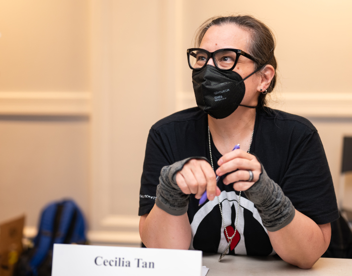 A small author with large glasses and a black KN94 mask, holding a pen, looking up at someone who is asking for an autograph