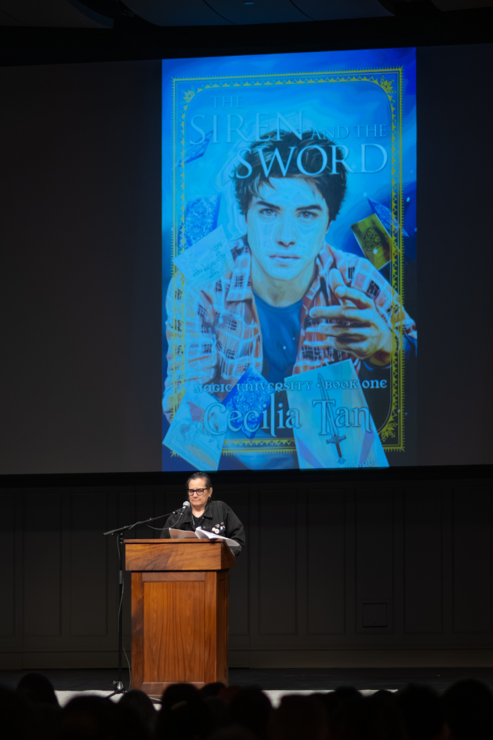 A small person at a large lectern with an even larger projection screen behind them. The book's title is The Siren and the Sword. Art by Fox Estacado.